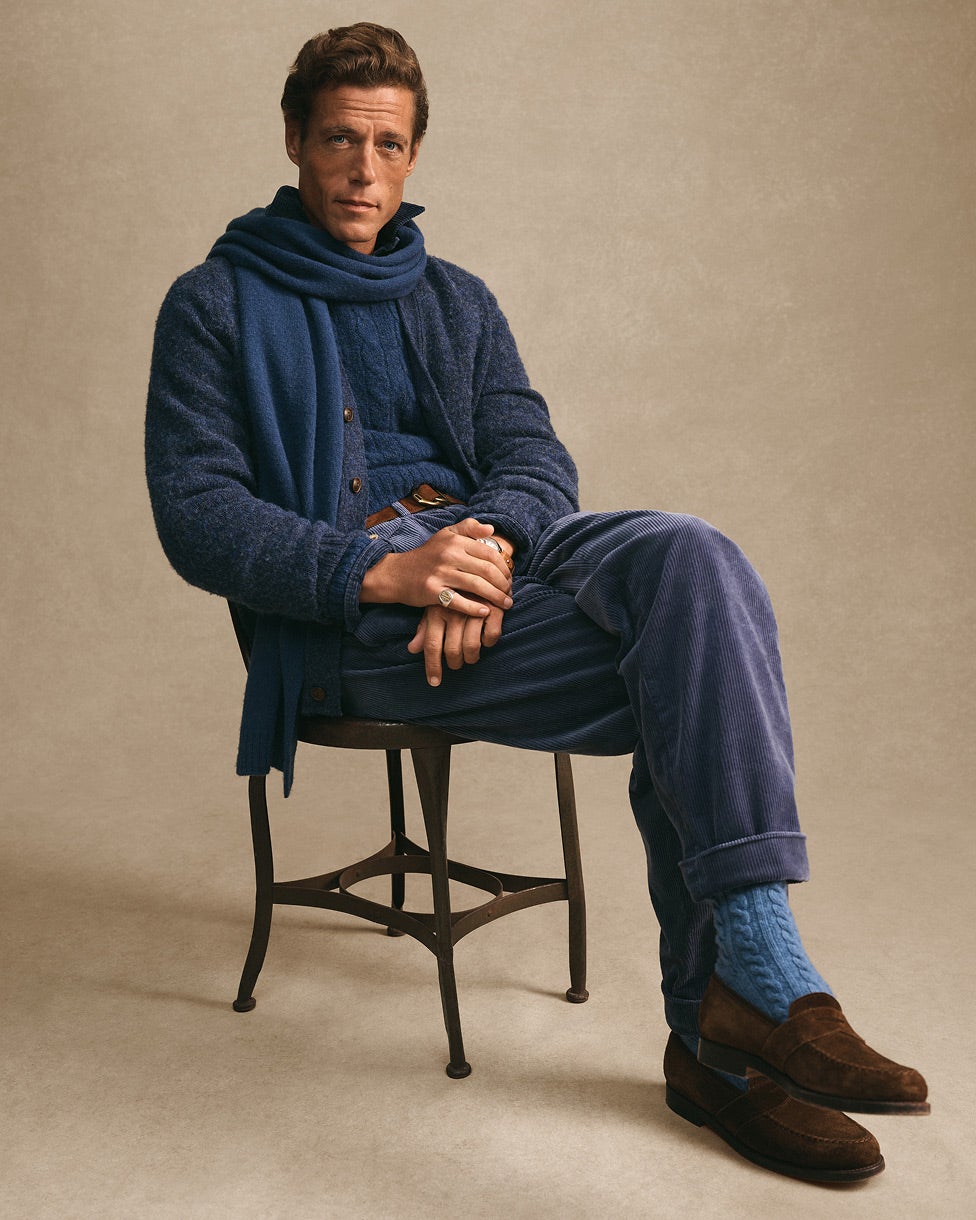 Hombre sentado vestido con ropa de punto azul, bufanda a juego y mocasines marrones.