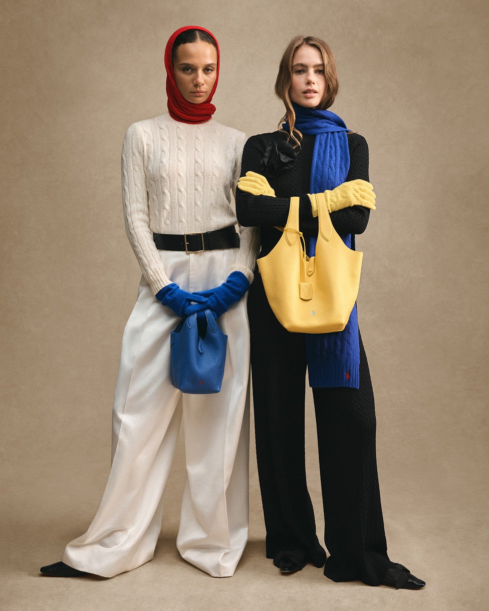 Dos mujeres de pie, una con una prenda de punto de color crema, un pañuelo rojo en la cabeza y guantes azules, y la otra con una prenda de punto negra, una bufanda azul y un bolso amarillo.